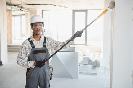Young african american construction worker painting walls using a telescopic paint roller in a...