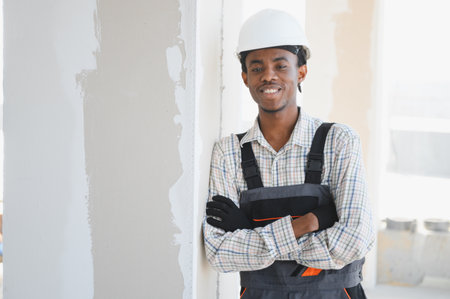 Young construction worker smiling with crossed arms, wearing a hardhat and work overalls, leaning...
