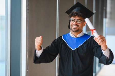 Excited graduate wearing graduation gown and cap holding diploma and celebrating successful...
