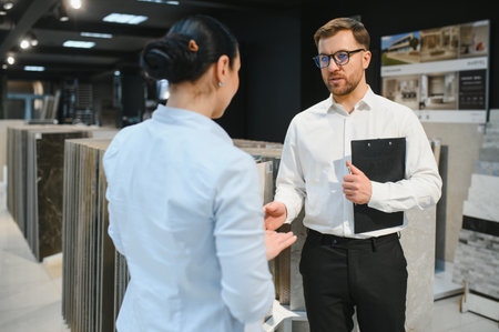 Shop assistant holding clipboard showing ceramic tiles to woman in hardware store, she is choosing material for her renovation projectの写真素材