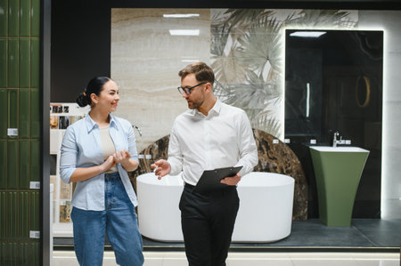 Salesman engaging in conversation with female customer while discussing various bathroom fixtures in a modern ceramic tile and plumbing storeの写真素材