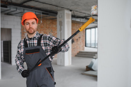 Construction worker wearing an orange helmet and overalls painting walls with a long roller in a building under constructionの写真素材