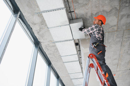 Construction worker installing an air conditioning duct on a ceiling, using a ladder in a building under constructionの写真素材