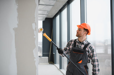 Professional builder using a paint roller, applying a fresh coat of paint to the walls of a building under renovation, demonstrating the process of interior finishingの写真素材