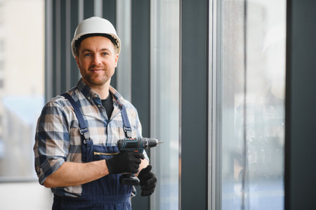 Construction worker wearing safety helmet and holding cordless drill while installing windows in a...