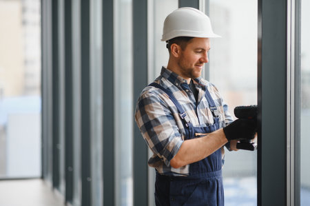 Construction worker wearing safety helmet and using cordless screwdriver while installing windows in a modern buildingの写真素材