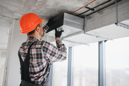 Builder wearing safety helmet is working on air conditioning and ventilation system installation in a building under constructionの写真素材