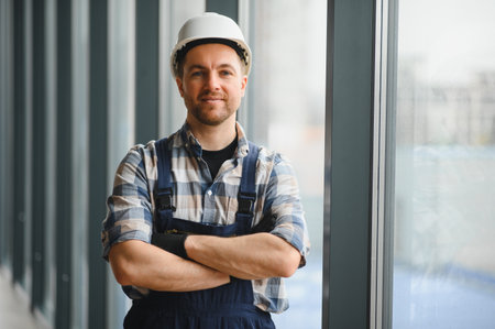 Portrait of smiling hvac technician wearing hardhat and work uniform standing with crossed arms by window in modern buildingの写真素材
