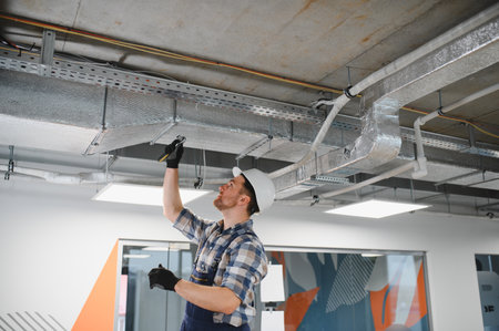 Hvac technician wearing safety helmet and work overalls installing an air conditioning system in an office buildingの写真素材