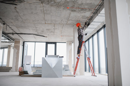 Electrician working on ventilation in an unfinished building, using a ladder and wearing safety equipmentの写真素材