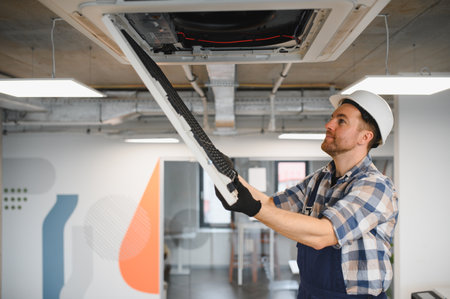 Hvac technician wearing safety helmet and gloves repairing cassette type air conditioning unit in modern office buildingの写真素材