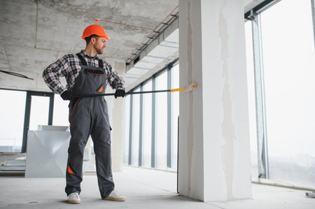 Construction worker wearing protective helmet and overalls, painting a wall using a long roller,...