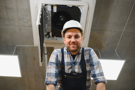 Hvac technician wearing hardhat and smiling while repairing ceiling mounted air conditioner unitの写真素材