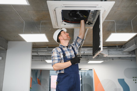 Hvac technician wearing safety helmet and gloves, repairing ceiling mounted cassette type air...