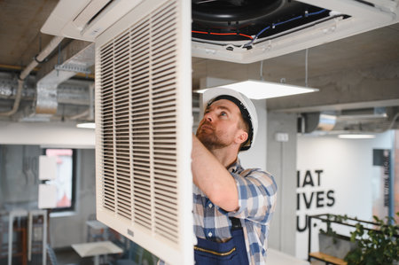 Hvac technician wearing safety helmet repairing ceiling mounted air conditioning unit in office buildingの写真素材