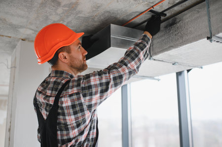 Skilled builder in a hard hat adjusting air conditioning ventilation ducts on a construction site,...