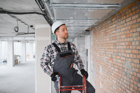 Construction worker wearing safety helmet and work overalls inspecting ventilation system while standing on a ladder in a building under constructionの写真素材