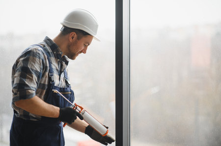 Construction worker wearing protective helmet and gloves applying sealant to a window frame during...