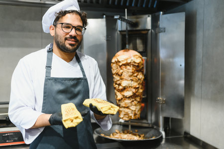 Cook wearing black gloves and apron, holding two shawarmas in a restaurant kitchen, next to a...