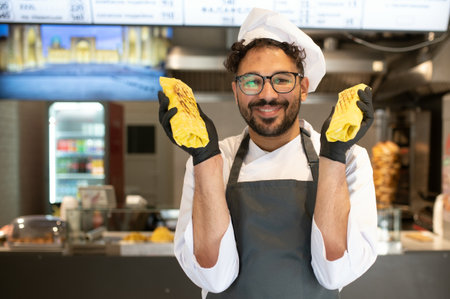 Happy cook showing two delicious shawarmas, wearing black gloves and a toque, working in his street...