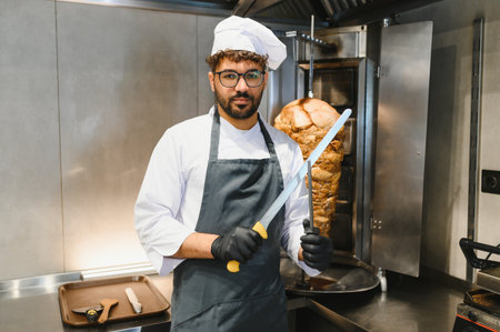 Professional chef holding knives and posing in front of a rotating shawarma spit in a commercial kitchen, ready to prepare delicious street foodの写真素材