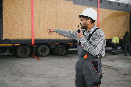 Construction worker wearing safety helmet and using walkie talkie while directing modular house...