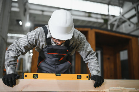 Focused carpenter using spirit level ensuring perfect alignment of wooden plank, demonstrating precision and quality craftsmanship in prefabricated house constructionの写真素材