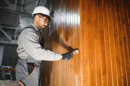 Focused carpenter varnishing prefabricated wooden wall section in modular house construction...