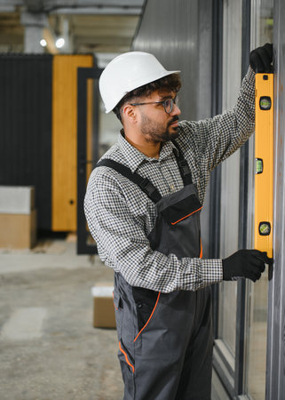 Focused construction worker checking vertical alignment of modular house wall using spirit level,...