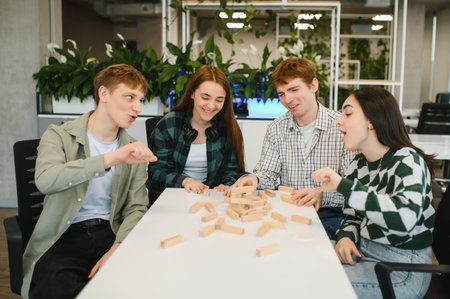 Four cheerful college students are concentrating while playing a wooden block game in a modern...