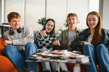 Four cheerful young people are sitting on beanbag chairs around a table, playing cards and having...