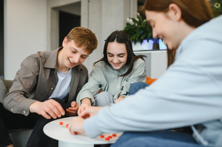 Three cheerful teenagers enjoying their leisure time, playing a board game together in a cozy home...