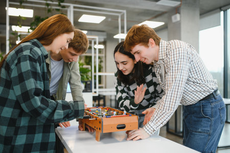 Four cheerful young startup employees are playing table football in a modern office during their...