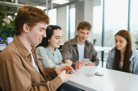 Four cheerful colleagues are playing cards at a table in a modern office, enjoying their break time...
