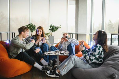 Four cheerful teenagers are sitting on comfortable beanbag chairs, playing cards and having fun together in a modern living roomの写真素材
