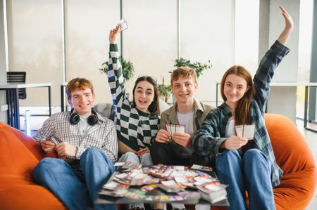 Four cheerful young students enjoying a lively card game at home, sharing laughter and making...