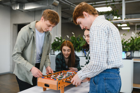 Four cheerful young startup employees are playing table football in a modern office, enjoying a fun...