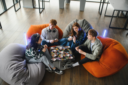 Four young startup employees are relaxing on beanbag chairs, playing a board game and having fun...