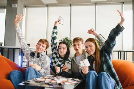 Four cheerful young students are sitting on an orange sofa, playing cards and having fun together