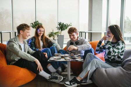 Four cheerful university students are sitting on beanbag chairs, playing cards and having fun in a...