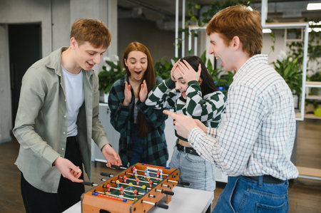 Four cheerful young office workers are enjoying their break time playing table football, showing excitement and having fun togetherの写真素材