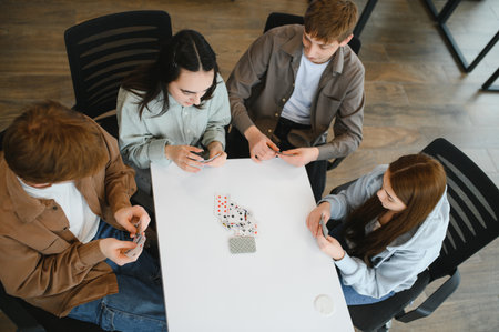 Four friends are sitting around a white table, playing cards and having fun, viewed from above