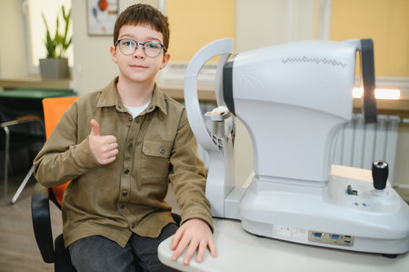 Young boy with glasses showing thumbs up during vision diagnostics with autorefractor in...