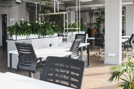 Modern office space with white desks, ergonomic chairs, and abundant potted plants creating a...