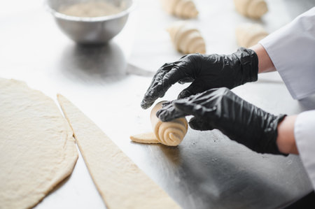 Baker wearing black gloves preparing croissants, rolling dough into shape on stainless steel table...