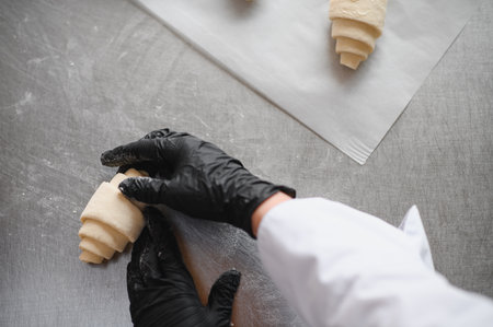 Baker wearing black gloves skillfully shapes croissant dough on a stainless steel table,...