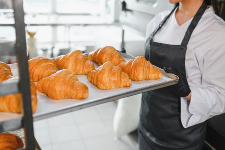 Baker wearing black gloves holding metal tray of warm, golden brown croissants in commercial kitchenの写真素材