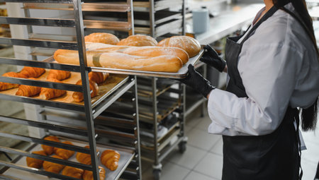 Baker wearing black gloves arranging freshly baked bread and croissants on metal rack in bakery...