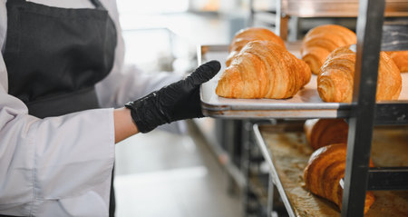 Baker wearing black gloves placing tray of golden brown croissants on metal rack in commercial...