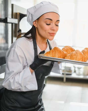 Professional baker woman smelling a tray of freshly baked golden croissants, enjoying the aroma in a...